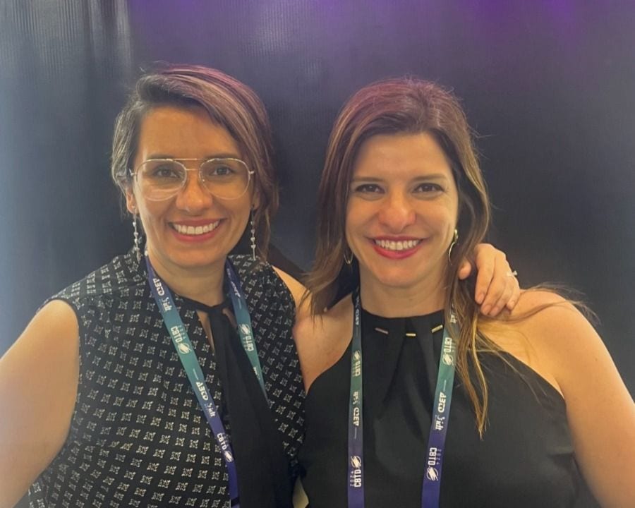 Duas mulheres estão de pé lado a lado, sorrindo, com crachás de conferência em volta do pescoço. Uma mulher está usando óculos e uma blusa estampada, enquanto a outra está usando uma blusa preta sem mangas.