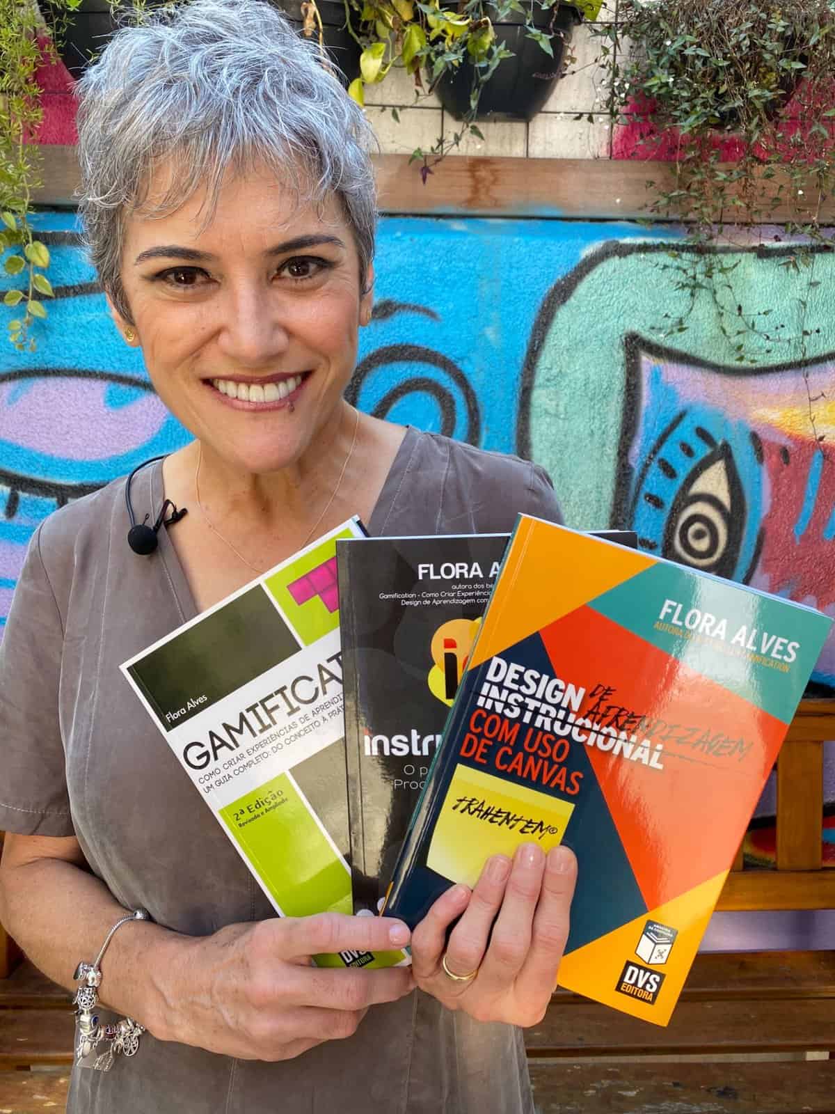 Uma pessoa com cabelos curtos e grisalhos sorri enquanto segura quatro livros. O fundo apresenta grafites coloridos.