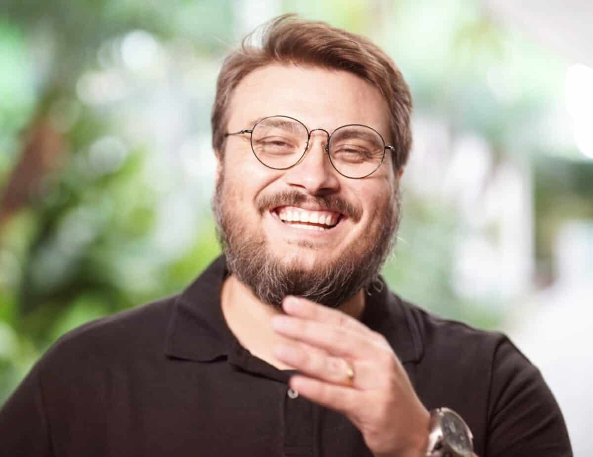 Um homem com óculos e barba está sorrindo largamente, vestindo uma camisa polo preta. Ele parece estar ao ar livre com vegetação desfocada ao fundo.