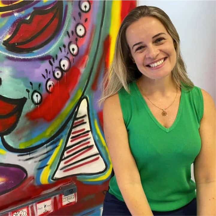 Uma mulher vestindo uma blusa verde sem mangas sorri em frente a um mural colorido e abstrato.