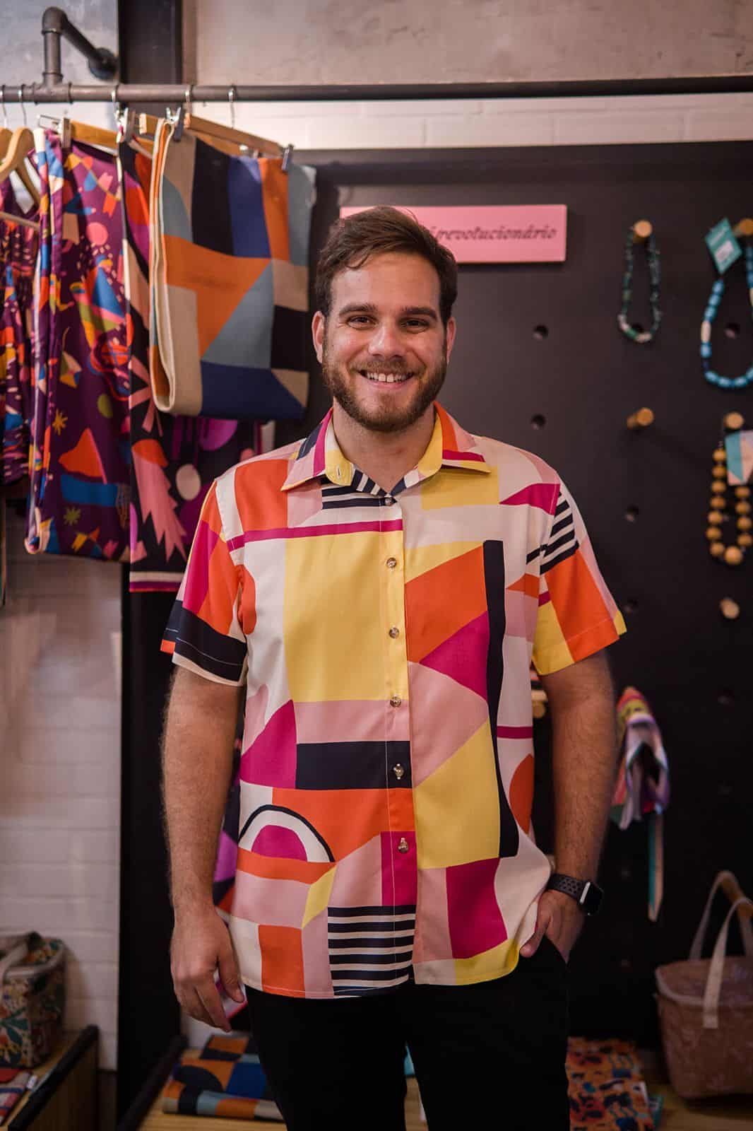 Uma pessoa vestindo uma camisa colorida com estampas geométricas está em frente a uma vitrine com roupas e acessórios de estilo semelhante.