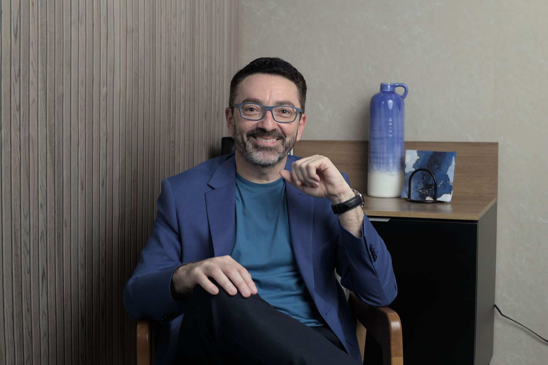 Um homem usando óculos e um blazer azul está sentado em uma cadeira de madeira em um escritório. Um jarro de cerâmica azul e branco repousa sobre um armário atrás dele.