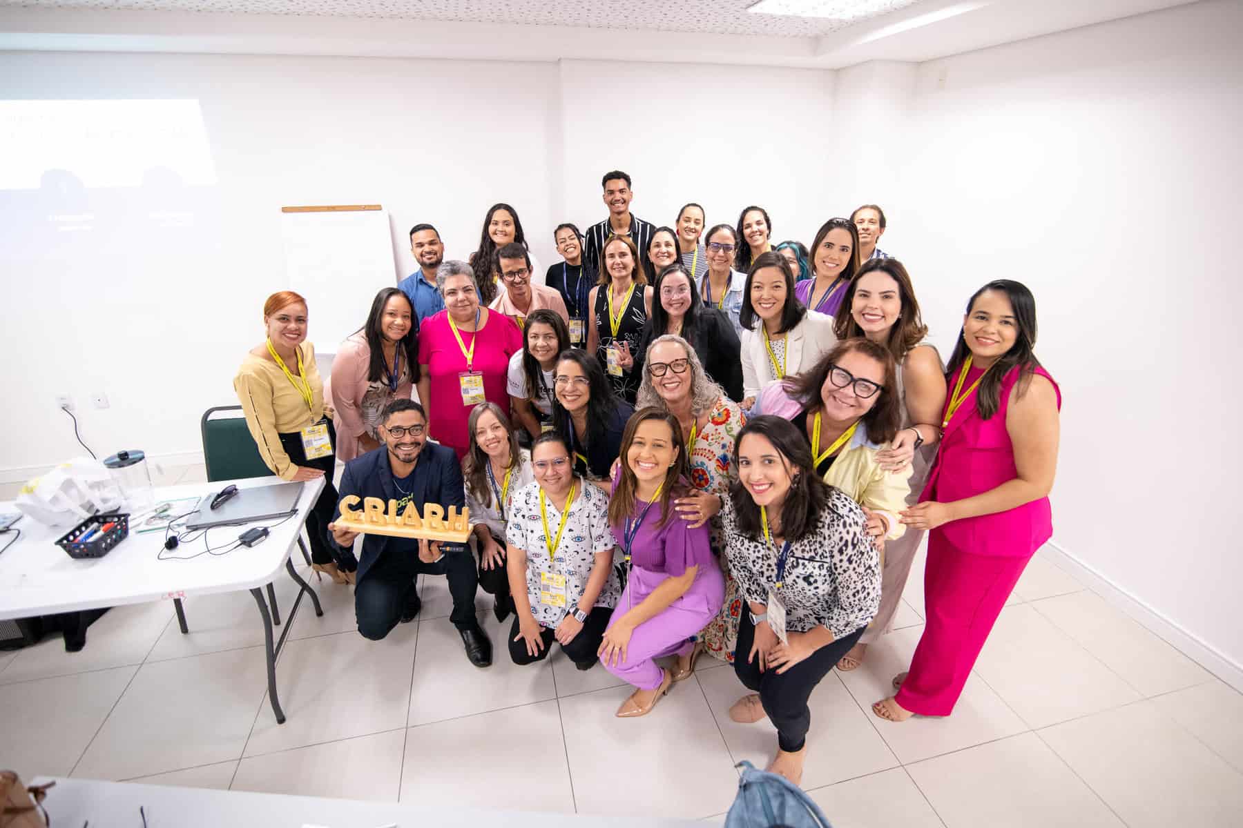 Um grupo de pessoas sorrindo e posando para uma foto de grupo em um ambiente de escritório. Alguns usam crachás e uma pessoa segura uma placa de madeira clara que diz “CRIAR.