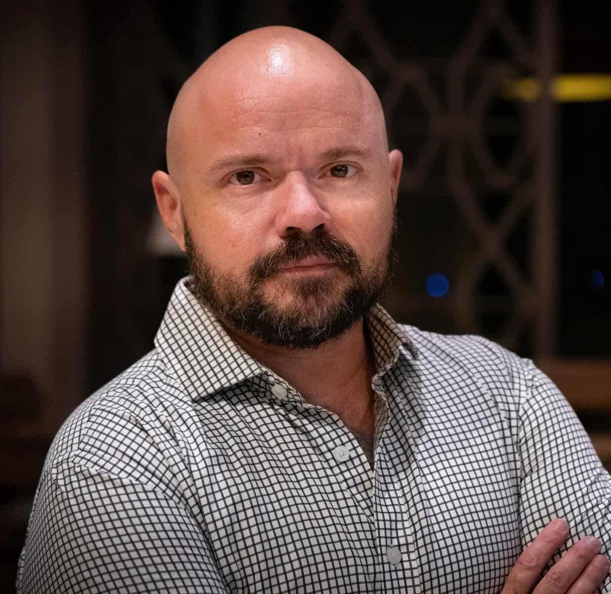 Um homem careca, com barba e bigode, vestindo uma camisa xadrez, está de braços cruzados em um ambiente interno.