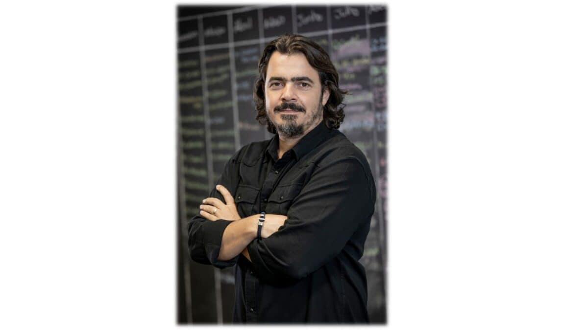 Um homem com cabelo de comprimento médio e barba está de braços cruzados, vestindo uma camisa preta, em frente a um quadro-negro coberto de escrita.