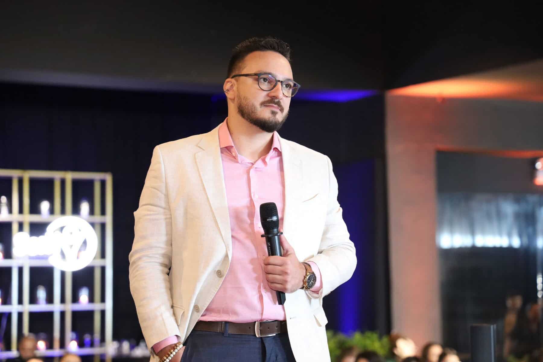 Um homem de blazer claro e óculos segura um microfone em pé em um ambiente interno, parecendo se dirigir a um público.