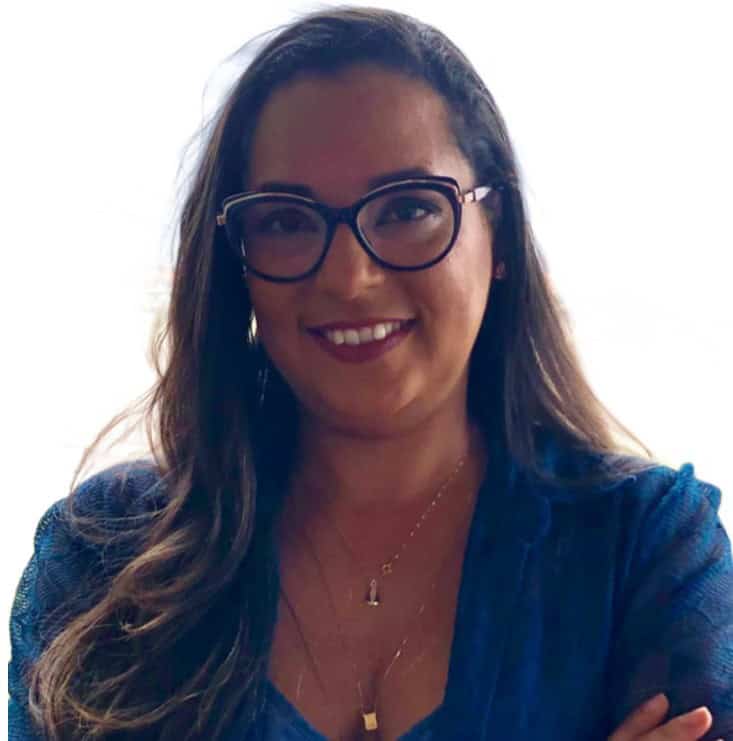 Uma mulher com longos cabelos castanhos e óculos, usando um top azul e colares em camadas, sorri para a câmera contra um fundo claro.