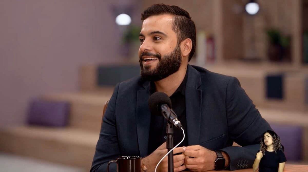 Um homem de terno escuro está sentado em uma mesa com um microfone, falando e sorrindo. Um intérprete de linguagem de sinais aparece no canto inferior direito.