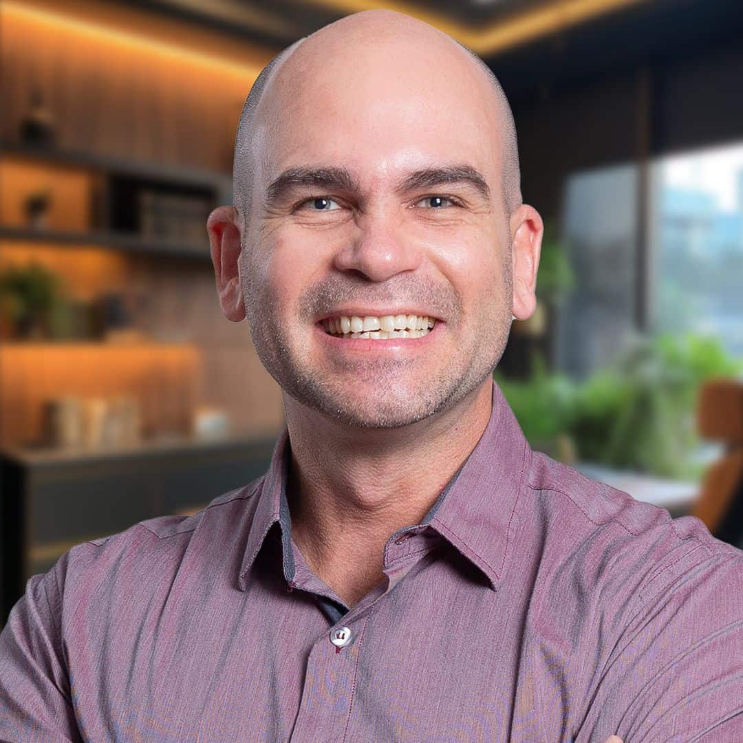 Um homem sorridente, careca, com uma camisa de botão lilás, em um escritório moderno com prateleiras e plantas ao fundo.