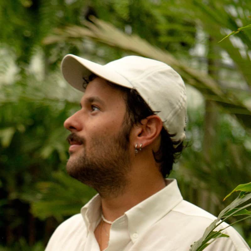 Um homem vestindo camisa branca e boné branco olha para a esquerda enquanto está em pé entre plantas verdes.