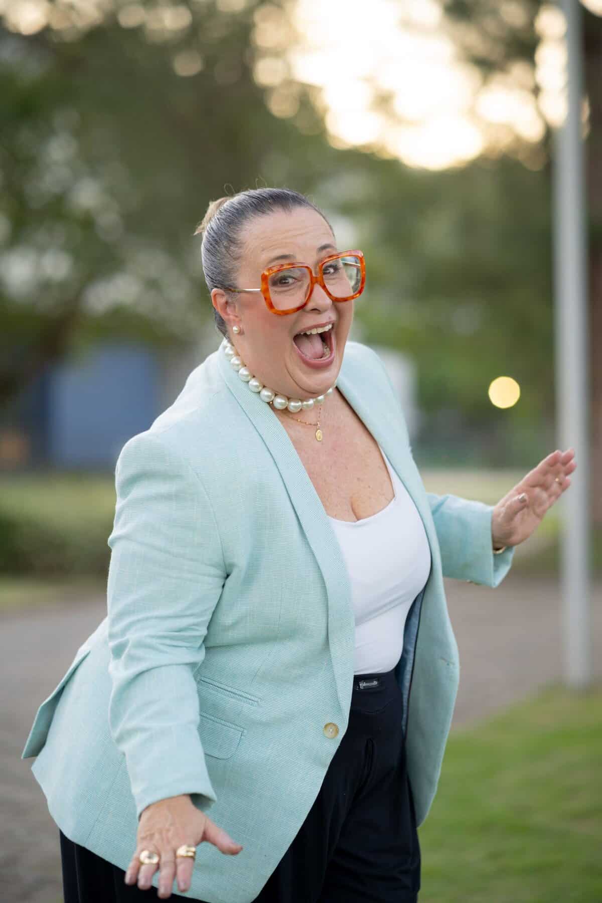 Uma mulher vestindo blazer azul-claro, blusa branca e grandes óculos laranja sorri e faz uma pose divertida ao ar livre em uma passarela.