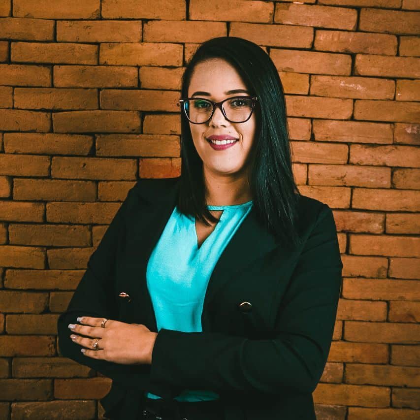 Uma mulher de cabelos escuros lisos e óculos sorri com os braços cruzados, parada em frente a uma parede de tijolos. Ela está vestindo um top azul-petróleo e um blazer preto.