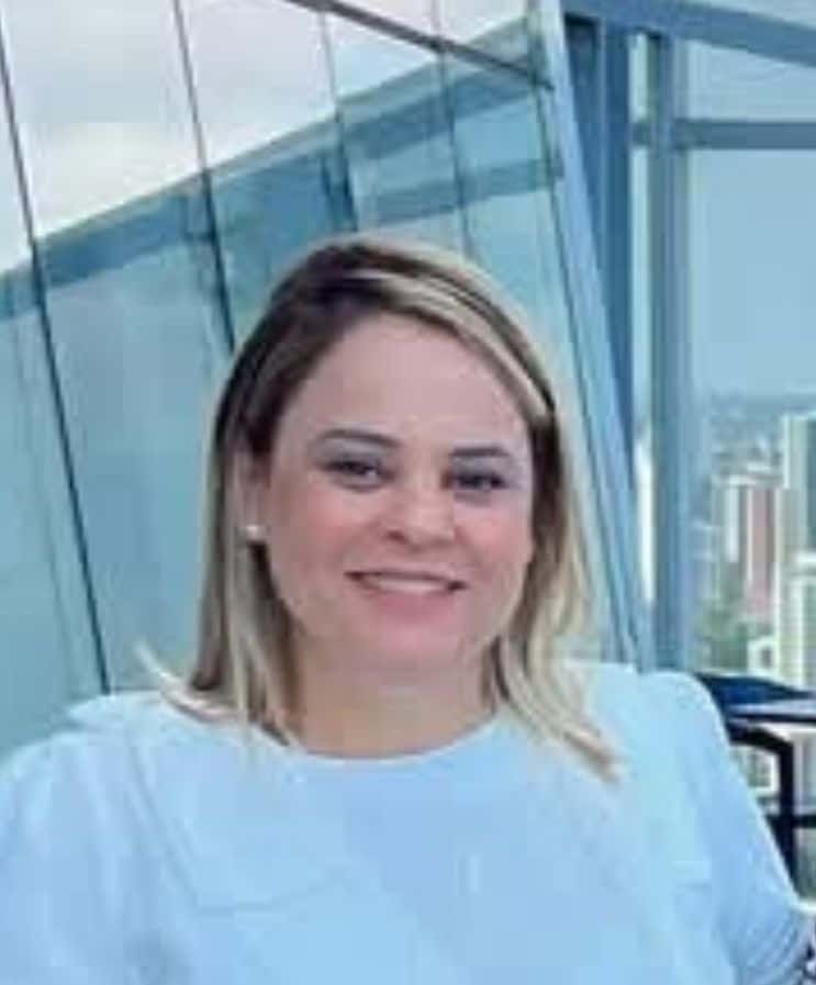 Uma mulher com cabelos loiros na altura dos ombros e um top azul claro sorri enquanto está em um ambiente interno próximo a grandes janelas com vista para uma paisagem urbana.