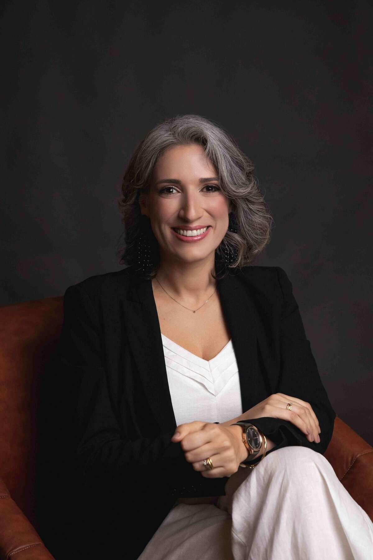 Uma mulher de cabelos grisalhos, vestindo um blazer preto e blusa branca, senta-se em uma cadeira marrom, sorrindo para a câmera contra um fundo escuro.