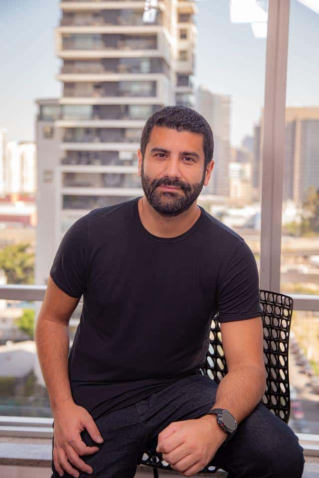 Um homem com barba e cabelo curto senta-se em uma cadeira em um ambiente interno, vestindo uma camiseta preta e um relógio, com os prédios da cidade visíveis pela janela atrás dele.