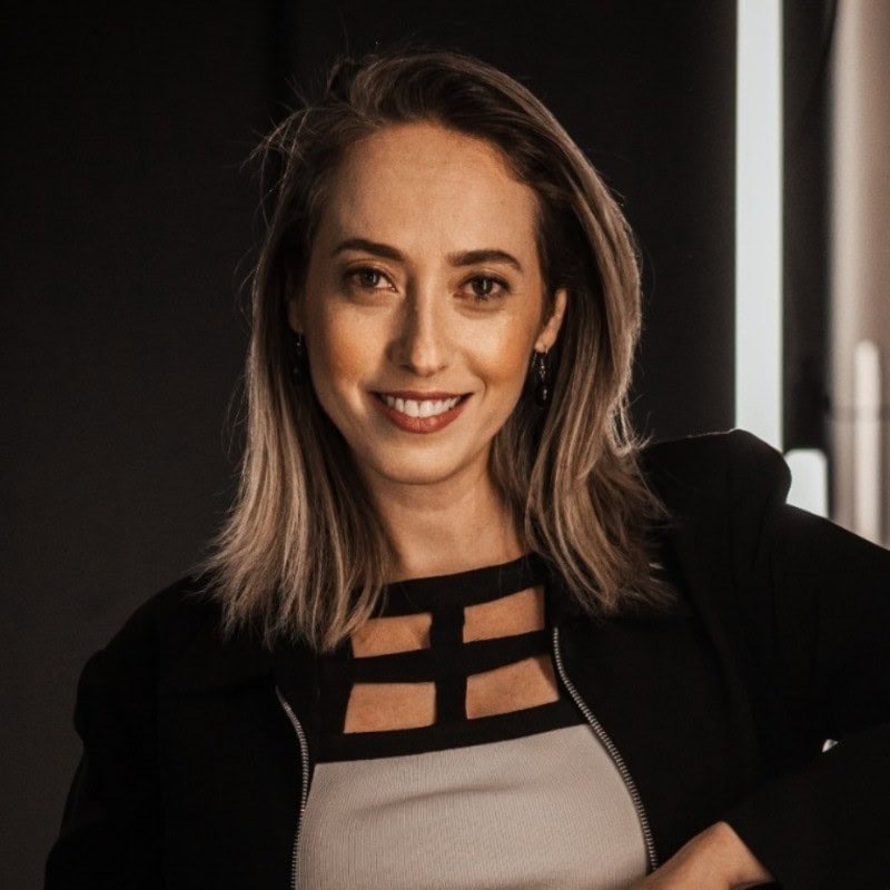 Uma mulher com cabelos loiros na altura dos ombros, vestindo um blazer preto e uma blusa estampada, sorrindo para a câmera contra um fundo escuro.