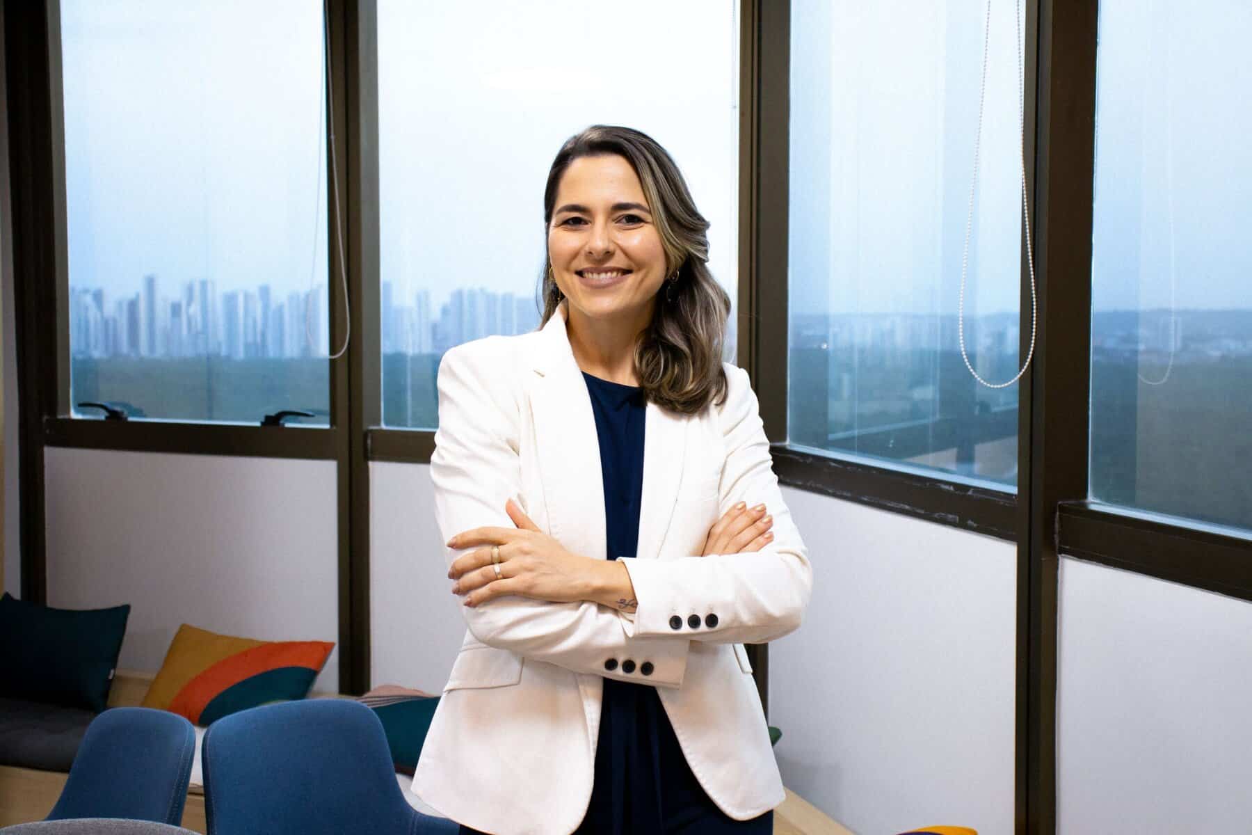 Uma mulher de blazer branco está de braços cruzados e sorri em um escritório moderno com grandes janelas e vista para a cidade.