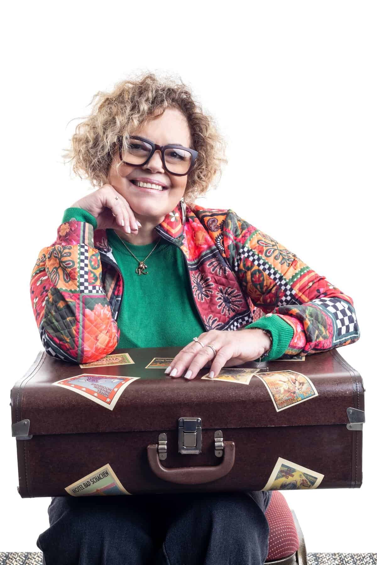 Uma pessoa sorridente com cabelos cacheados e óculos senta-se atrás de uma mala coberta de adesivos de viagem, vestindo uma jaqueta colorida e uma blusa verde contra um fundo branco.