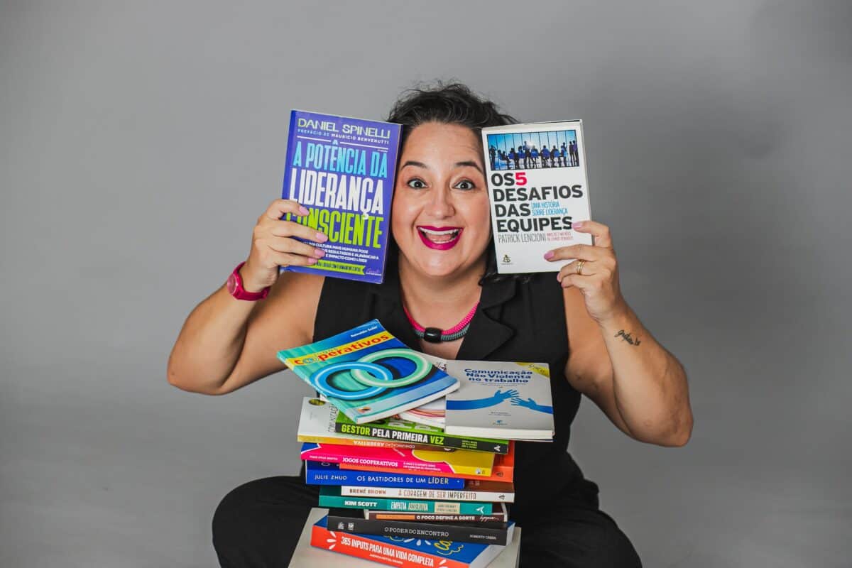 Uma pessoa sorridente está sentada atrás de uma pilha de livros, segurando dois livros sobre liderança e trabalho em equipe, em um fundo cinza claro.