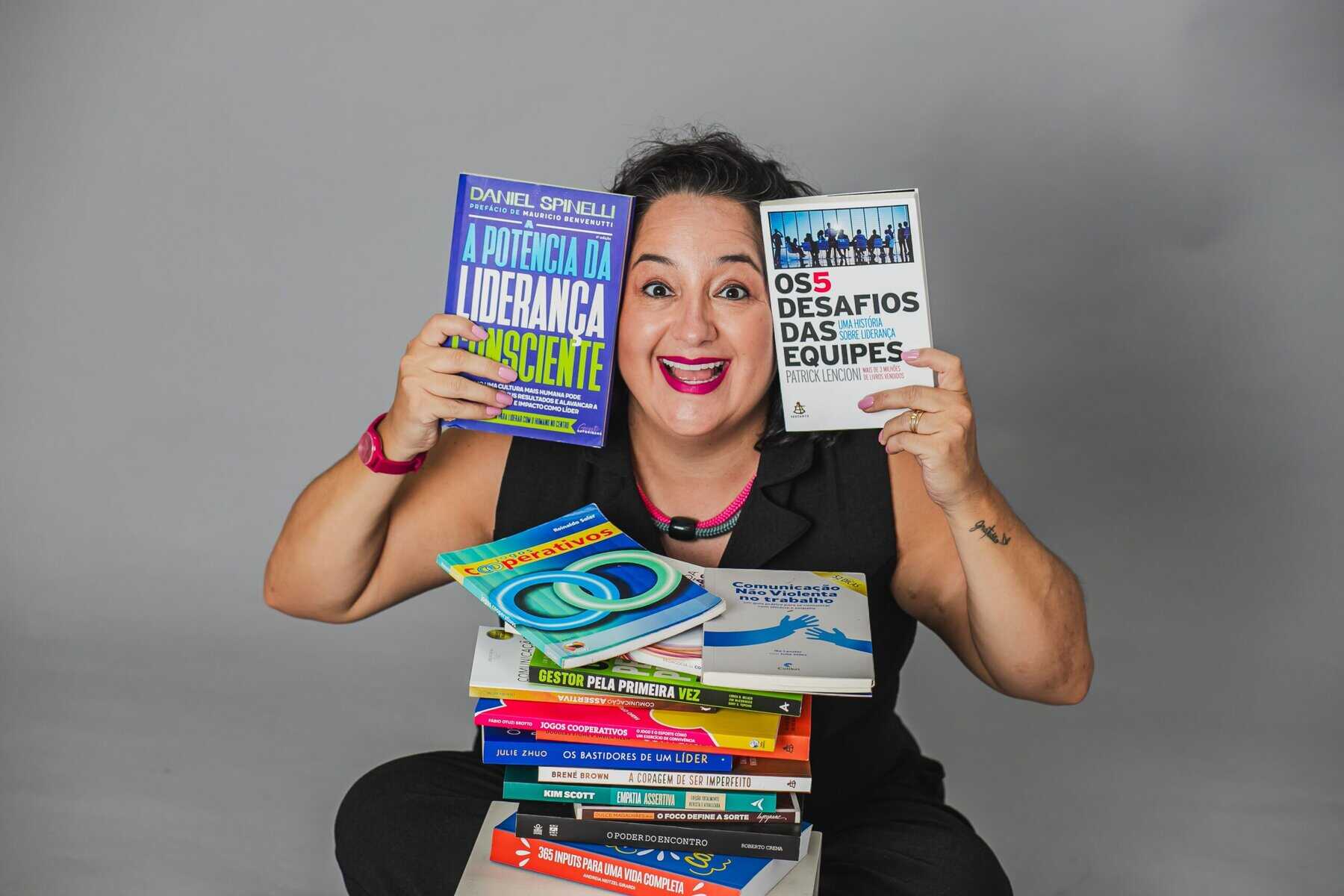 Uma pessoa sorridente está sentada atrás de uma pilha de livros, segurando dois livros sobre liderança e trabalho em equipe, em um fundo cinza claro.