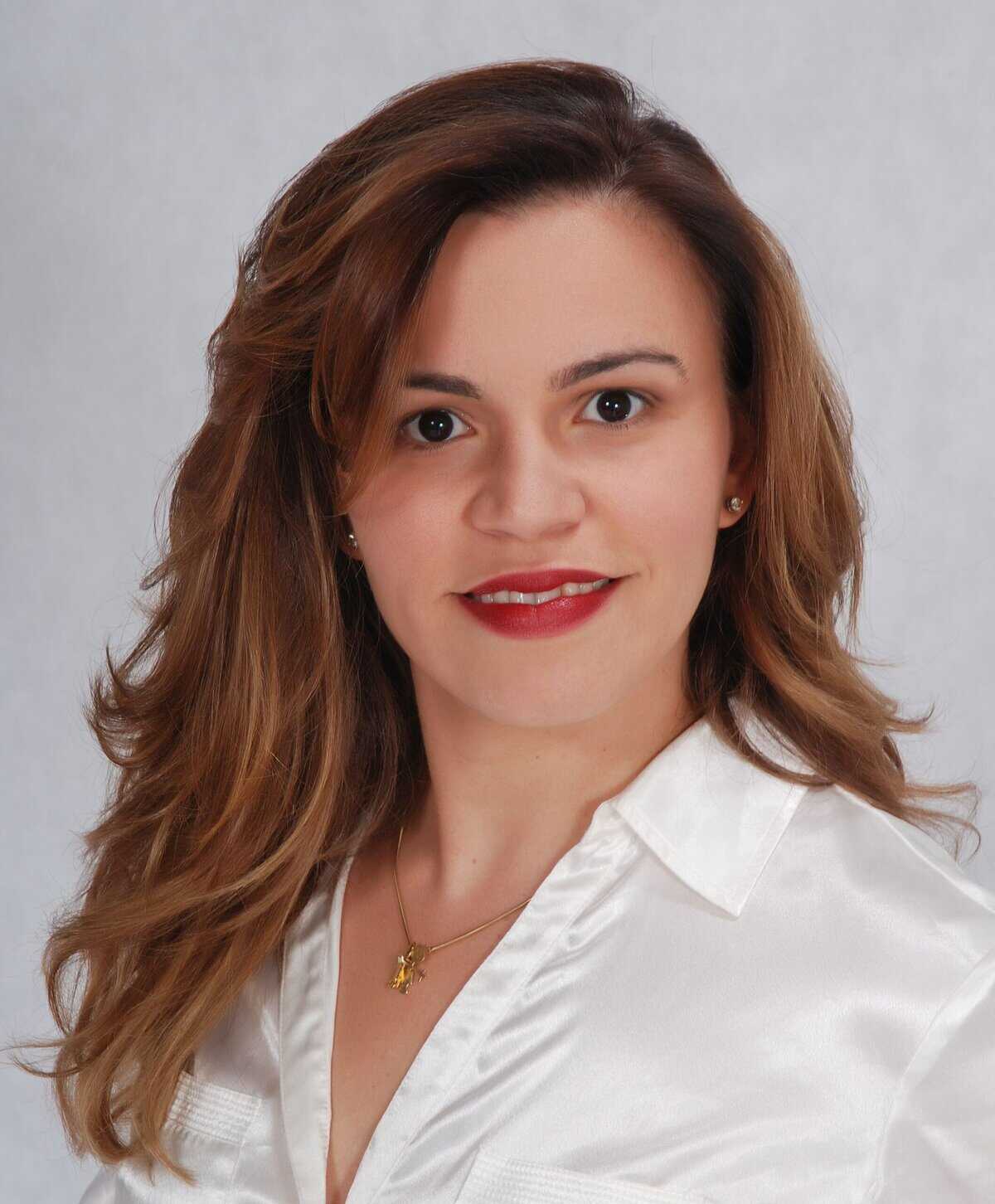 Mulher com longos cabelos castanhos, vestindo blusa branca e colar de ouro, posando em frente a um fundo claro e liso.
