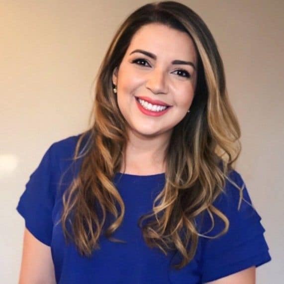 Mulher com longos cabelos castanhos ondulados e um top azul sorri para a câmera contra um fundo bege simples.